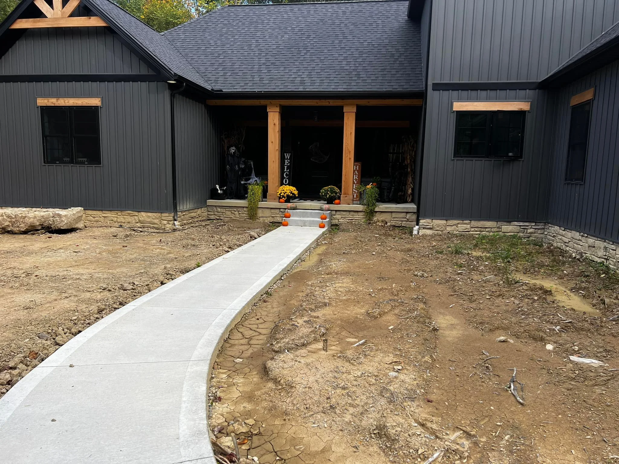 Front porch of a dark-colored house decorated with fall pumpkins, potted plants, and signs that say 'Welcome' and 'Harvest.' A concrete pathway leads to the porch, and the yard appears to be under landscaping or construction.