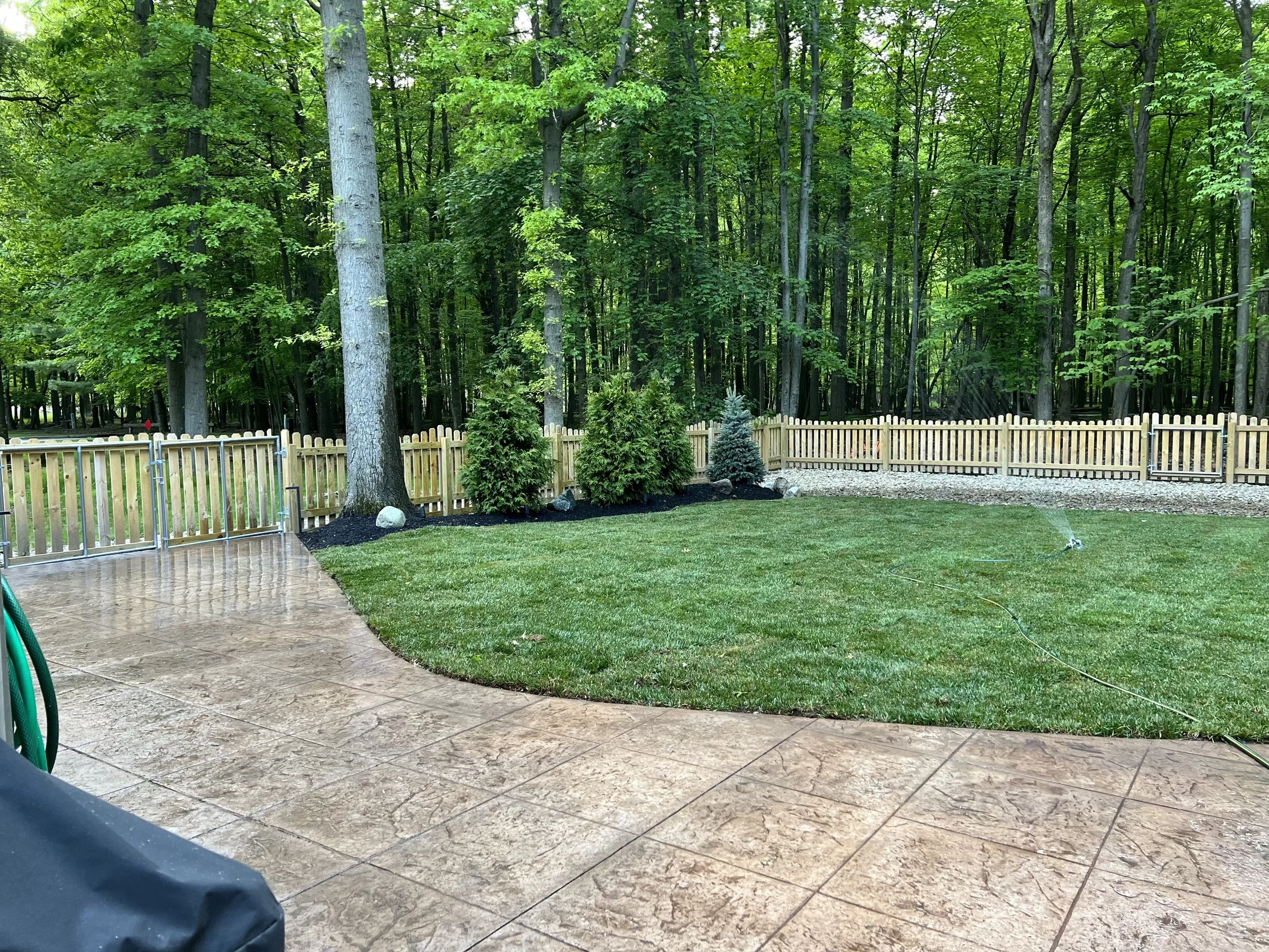 Backyard patio with a tan stamped concrete surface, a green lawn, a wooden picket fence, and a backdrop of tall trees in a wooded area.