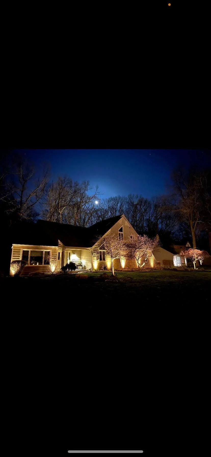 A house illuminated at night with outdoor lighting, surrounded by trees in bloom, under a clear sky with visible moon and stars.