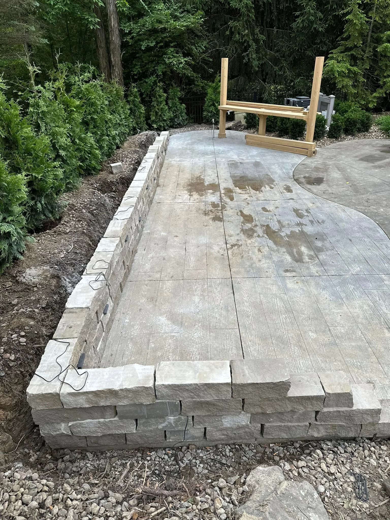 A backyard patio under construction with a partially built stone retaining wall and a concrete slab surface, surrounded by green bushes and trees.