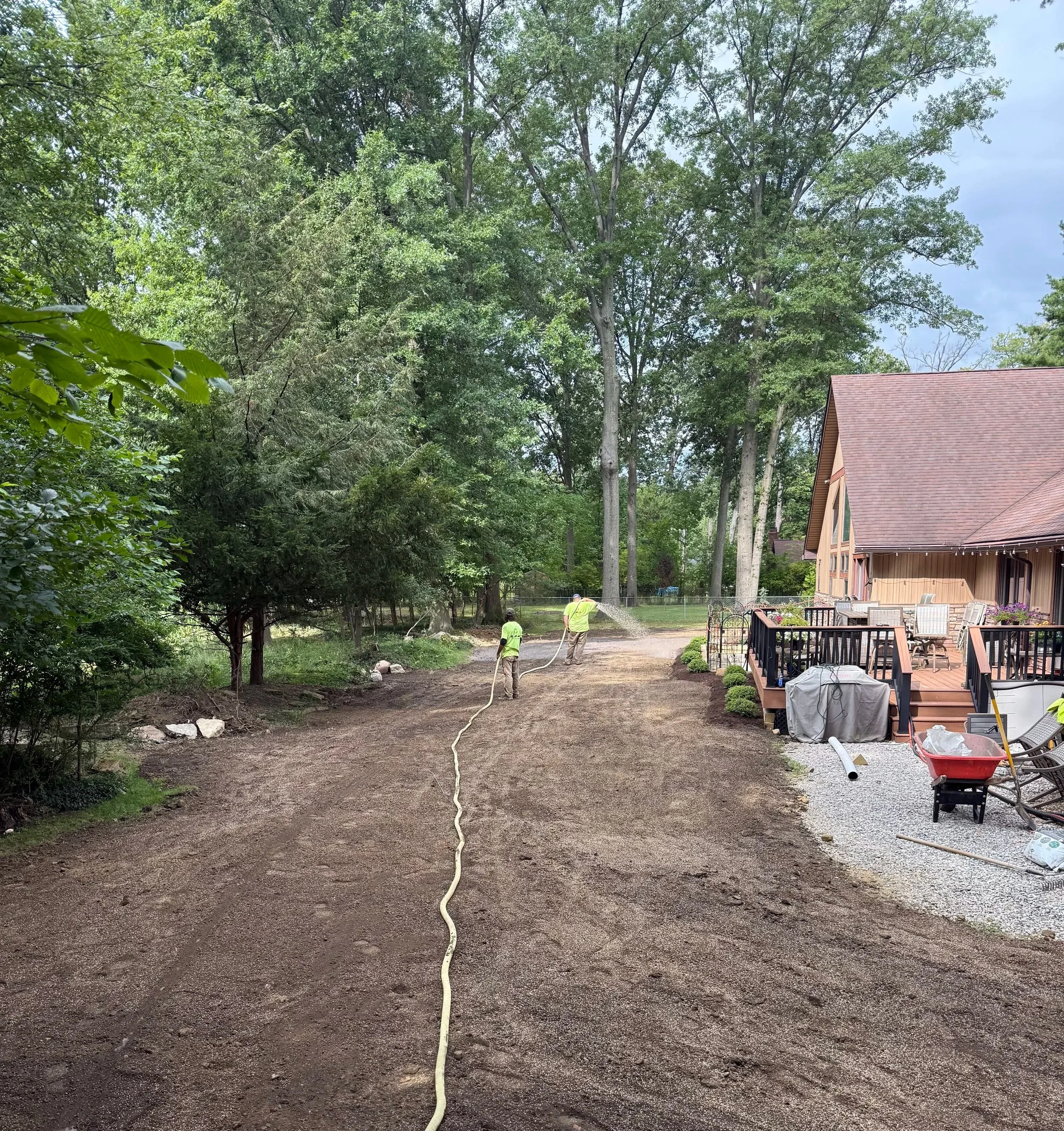 Three workers in neon yellow shirts are laying down a gravel driveway in a backyard, with a house on the right and tall trees in the background.
