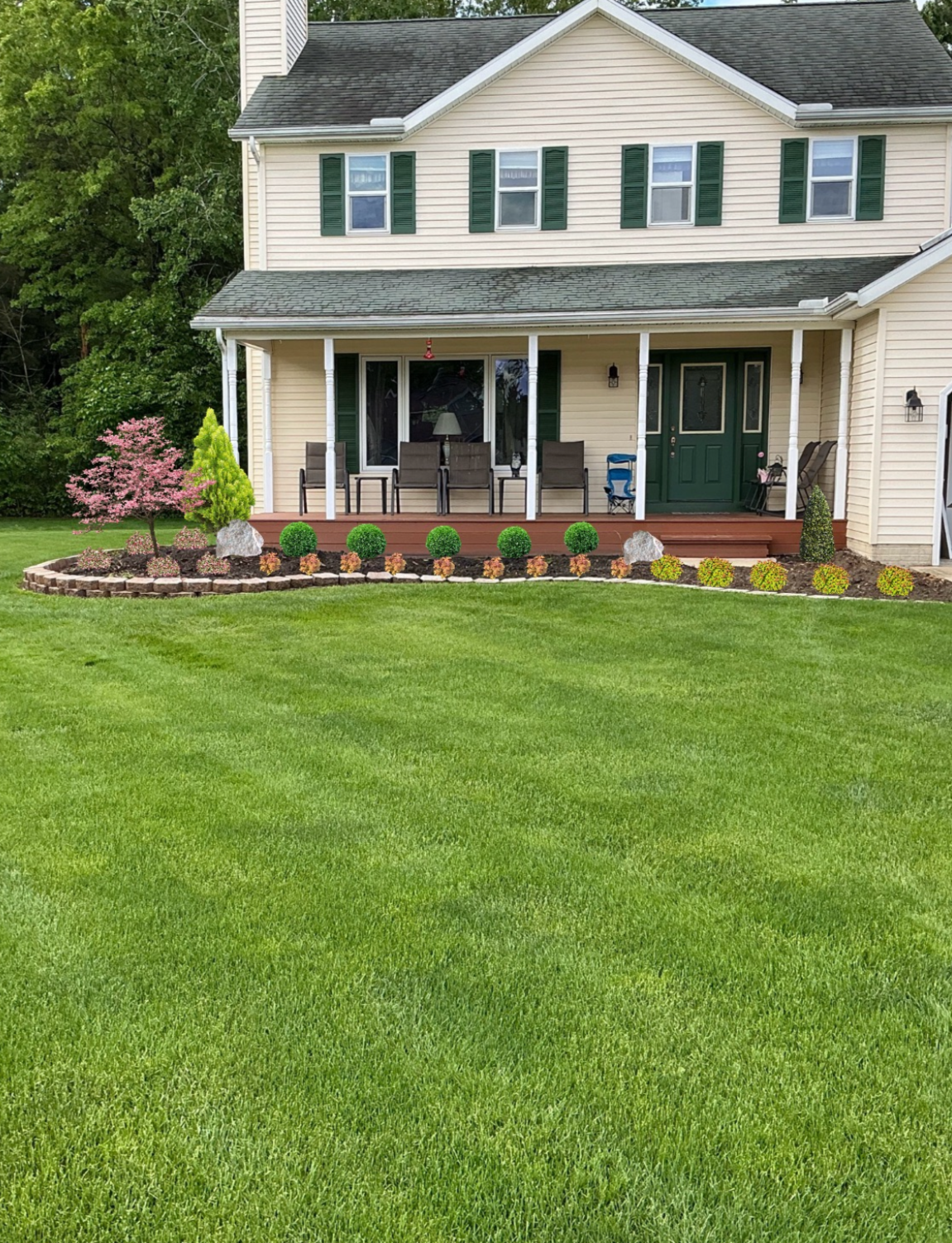 Rear view of a two-story house with a porch, green shutters, a green front door, and a well-maintained lawn with trimmed bushes and a small garden bed with colorful plants.