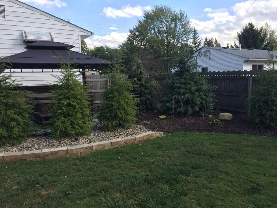 Backyard with a grassy lawn, a flower bed bordered with brick, small trees, and taller shrubs behind a wooden fence. There is a patio umbrella and a house with white siding in the background.