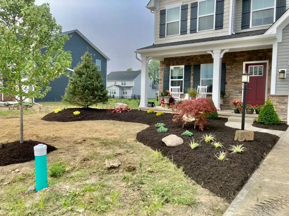 Front yard with a landscaped garden, small trees, and decorative plants in front of a house with porch and stone accents.