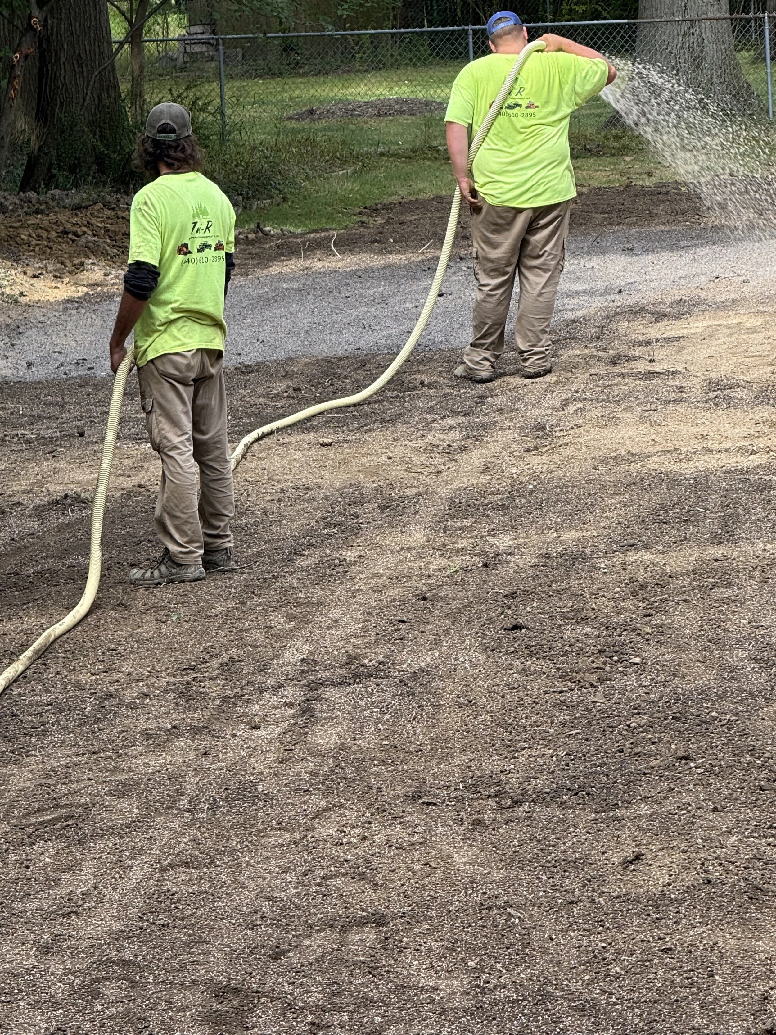 Two workers in neon green shirts are laying down a gravel or dirt surface in a yard, using a hose to spray water over the area.
