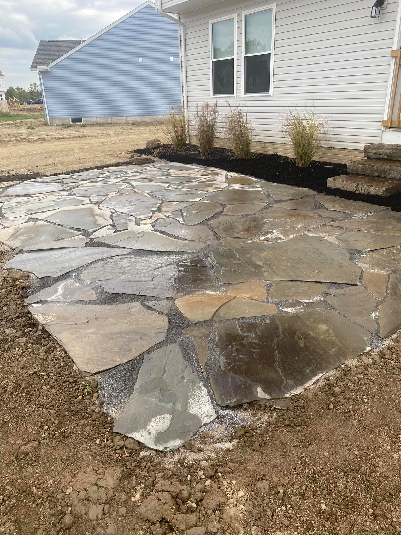 A newly installed flagstone patio with irregularly shaped stones in front of a house with white siding and a small set of stone steps. There are three ornamental grasses planted along the house foundation, and the ground around the patio is still dir