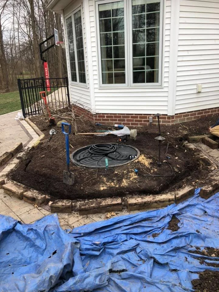 Front yard landscaping project with a circular brick border, soil preparation, gardening tools, and a black drainage basin, near a house with white siding and large windows.