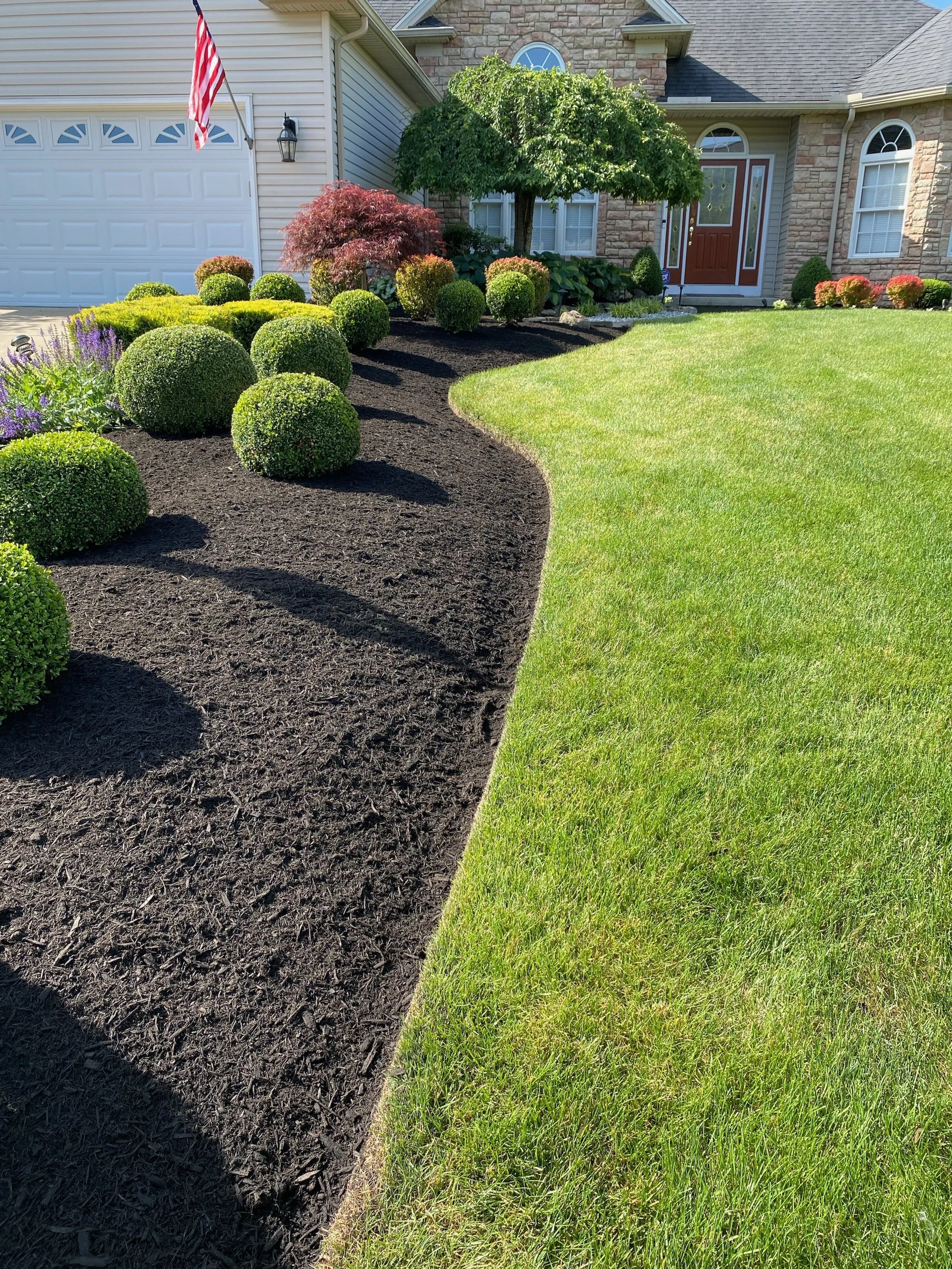 A well-maintained front yard with a green lawn, neatly trimmed bushes, and a curved garden bed with dark mulch, situated in front of a house with a stone and siding exterior and a red front door. An American flag is hanging near the garage.