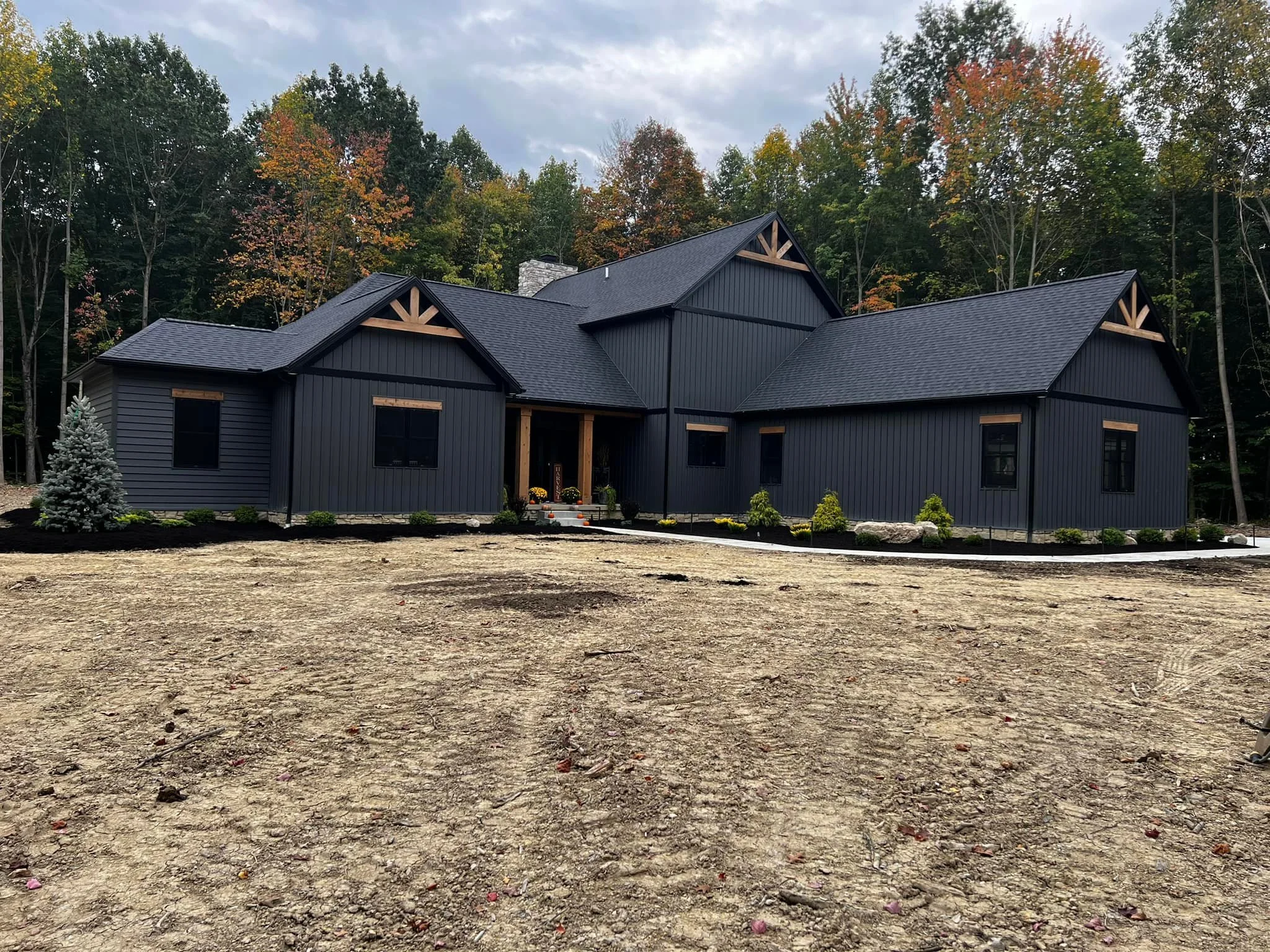 A modern dark gray house under construction or landscaping, with small plants and trees in the front yard, and a wooded area with trees showing fall colors in the background.