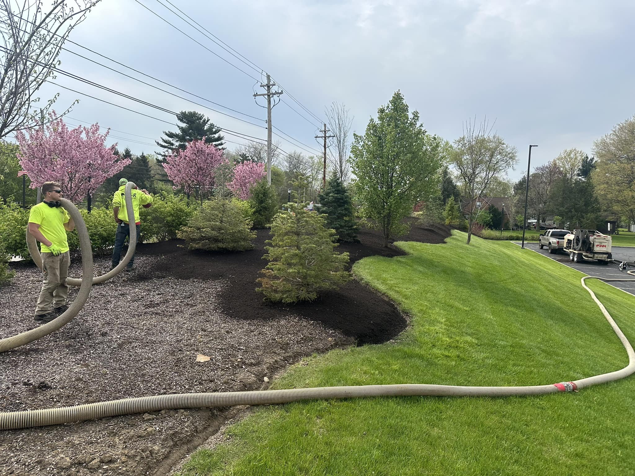 Workers using vacuum hoses to clean or perform maintenance on landscaped area with trees, grass, and flowering bushes in a parking lot area.