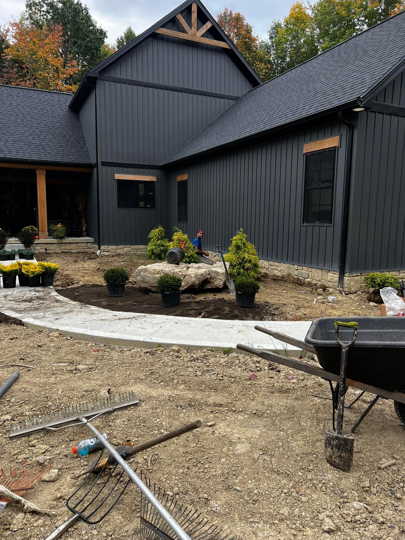 Construction worker sitting on a large rock in front of a new black building with landscaping plants and a sidewalk in progress.