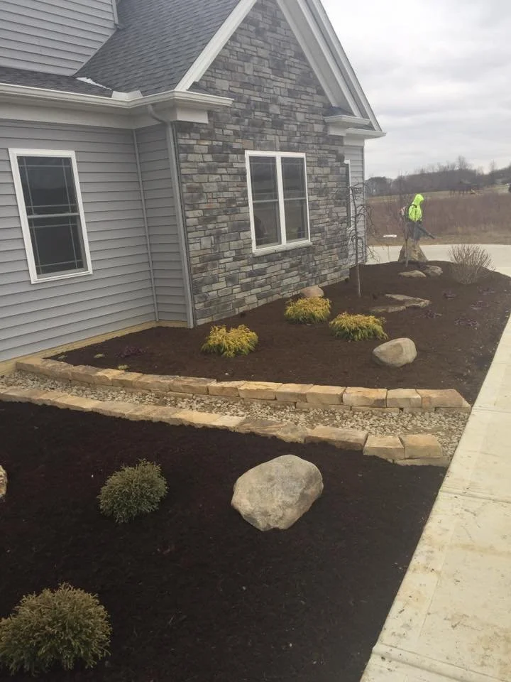 A newly landscaped front yard of a house featuring dark soil, small bushes, and large rocks, with a curved retaining wall made of bricks and a stone pathway. A worker in a bright yellow jacket is seen in the background.