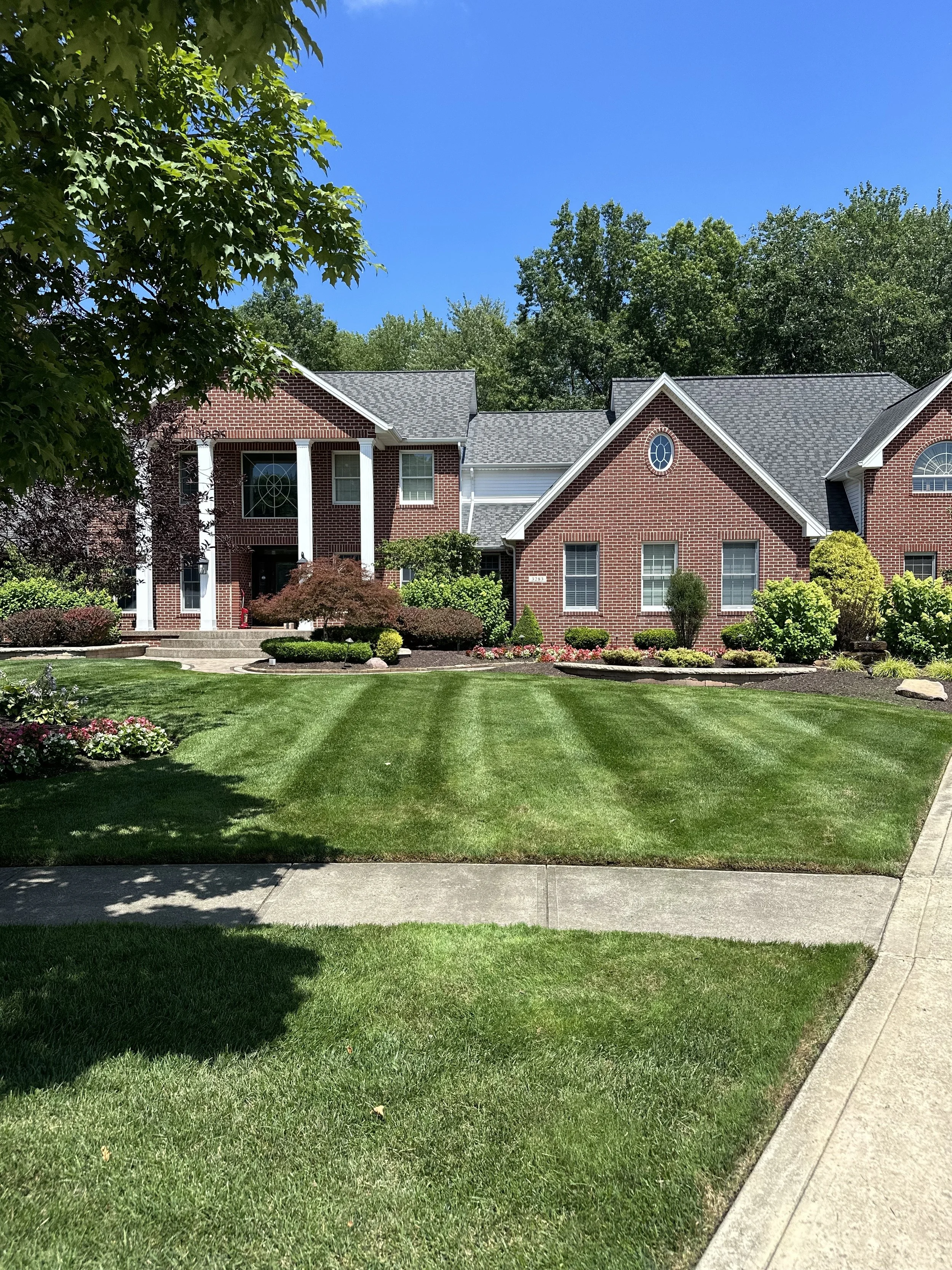 Front yard of a brick house with a well-maintained lawn, colorful flower beds, and a sidewalk, under a clear blue sky.