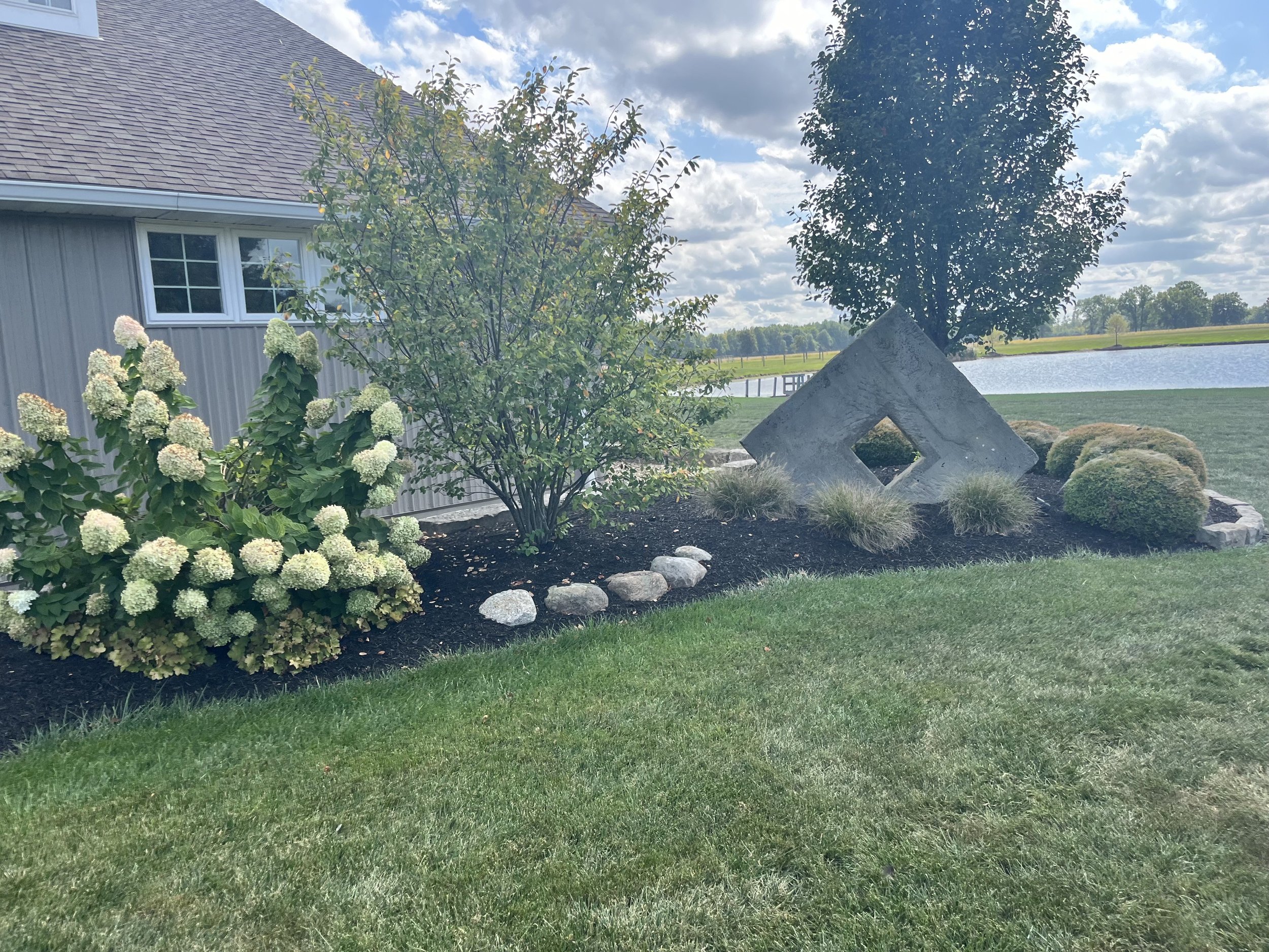 Landscaped yard with a garden bed next to a gray house, featuring blooming hydrangeas, a small tree, ornamental grass, and a concrete sculpture near a pond under a partly cloudy sky.