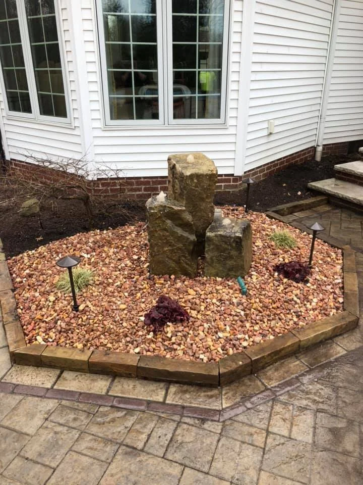 A small landscaped garden bed with pink gravel, a large decorative stone, two small plants, and two solar lights in front of a white house with vinyl siding and large windows.