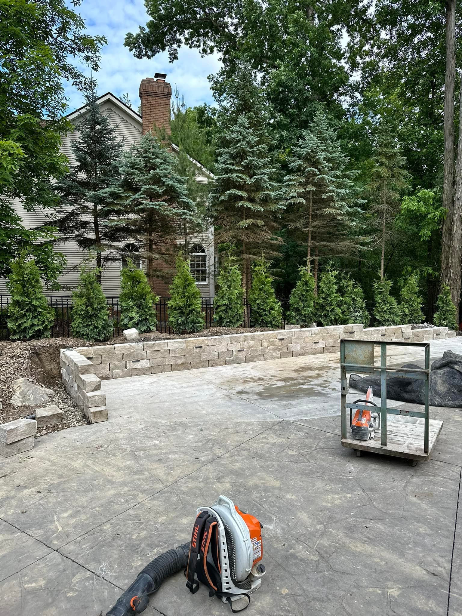 Construction site with a concrete patio, a stone retaining wall, and construction tools including a leaf blower and a cart, with trees and a house in the background.