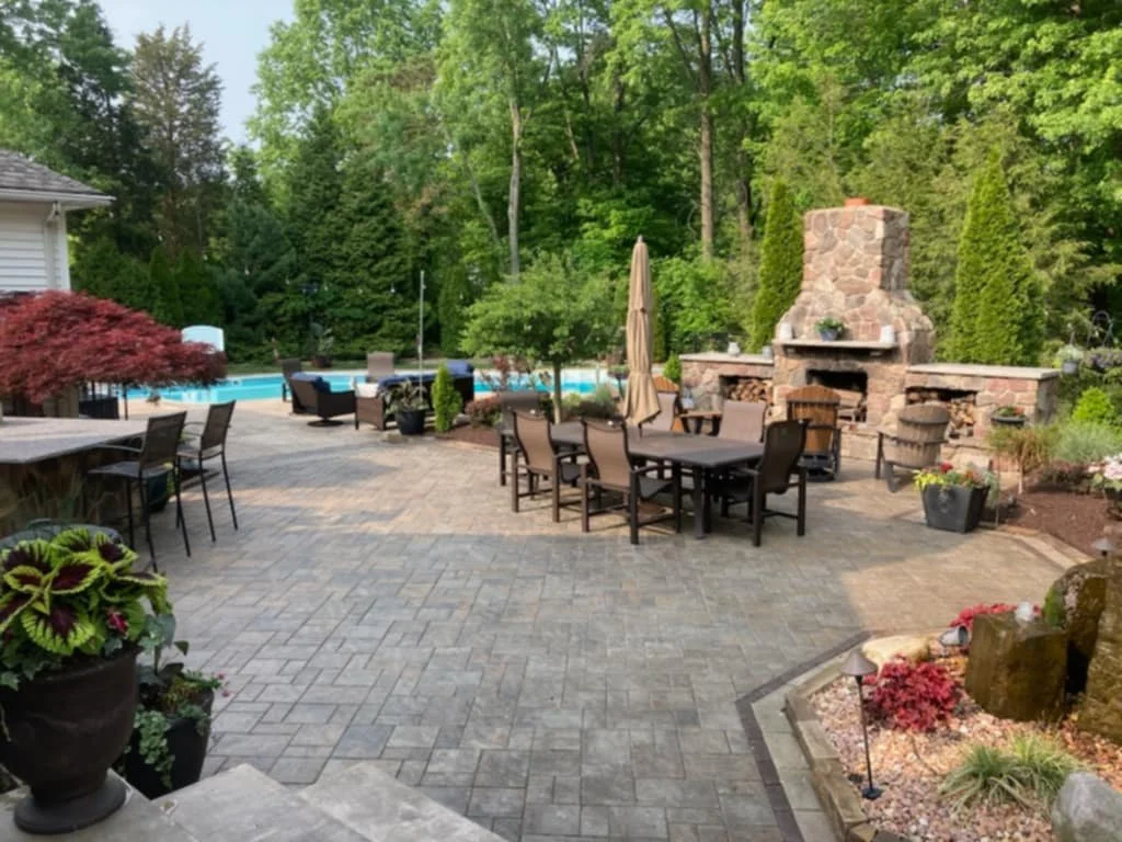 Backyard with a stone patio, outdoor dining furniture, a stone fireplace, a swimming pool, and lush green trees.