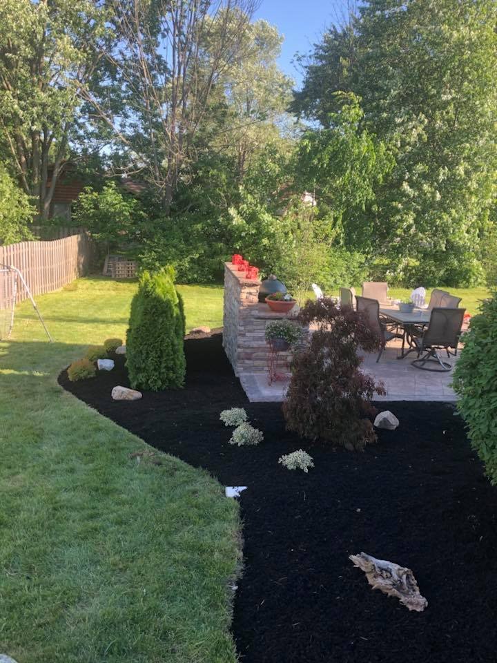 A backyard patio with a dining table and chairs, surrounded by lush green trees and a landscaped garden with freshly mulched beds, decorative rocks, and a brick wall.