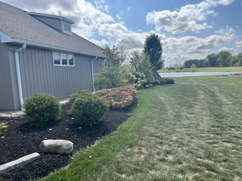 A well-maintained backyard with a house featuring gray siding, a manicured lawn, colorful shrubs and flowers, and a pond in the distance under a partly cloudy sky.
