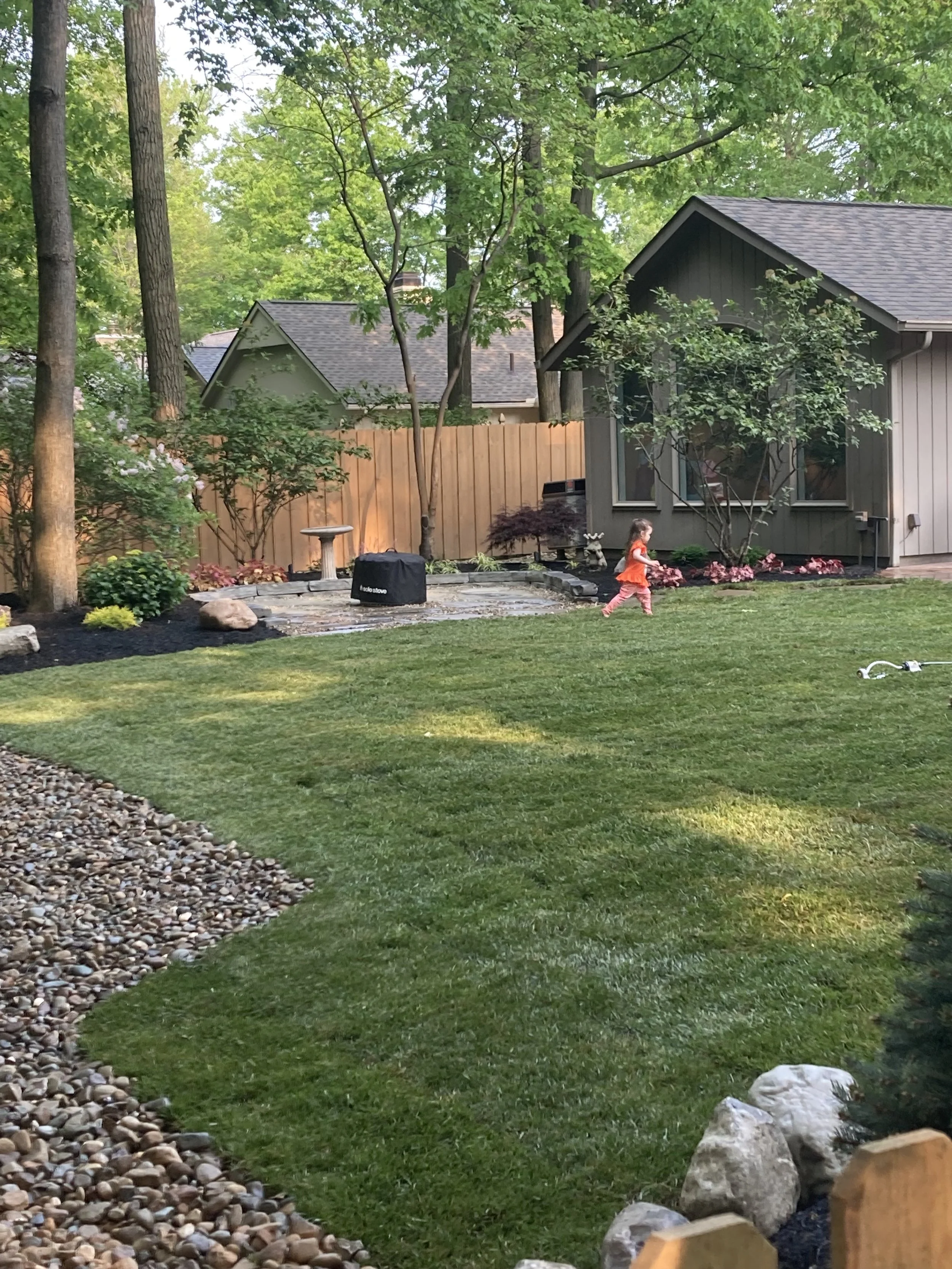 A backyard with green grass, trees, flowers, and a small child running. There is a house with gray siding and a dark roof, a wooden fence, and various plants along the fence line. A birdbath and a black object are visible on the patio.