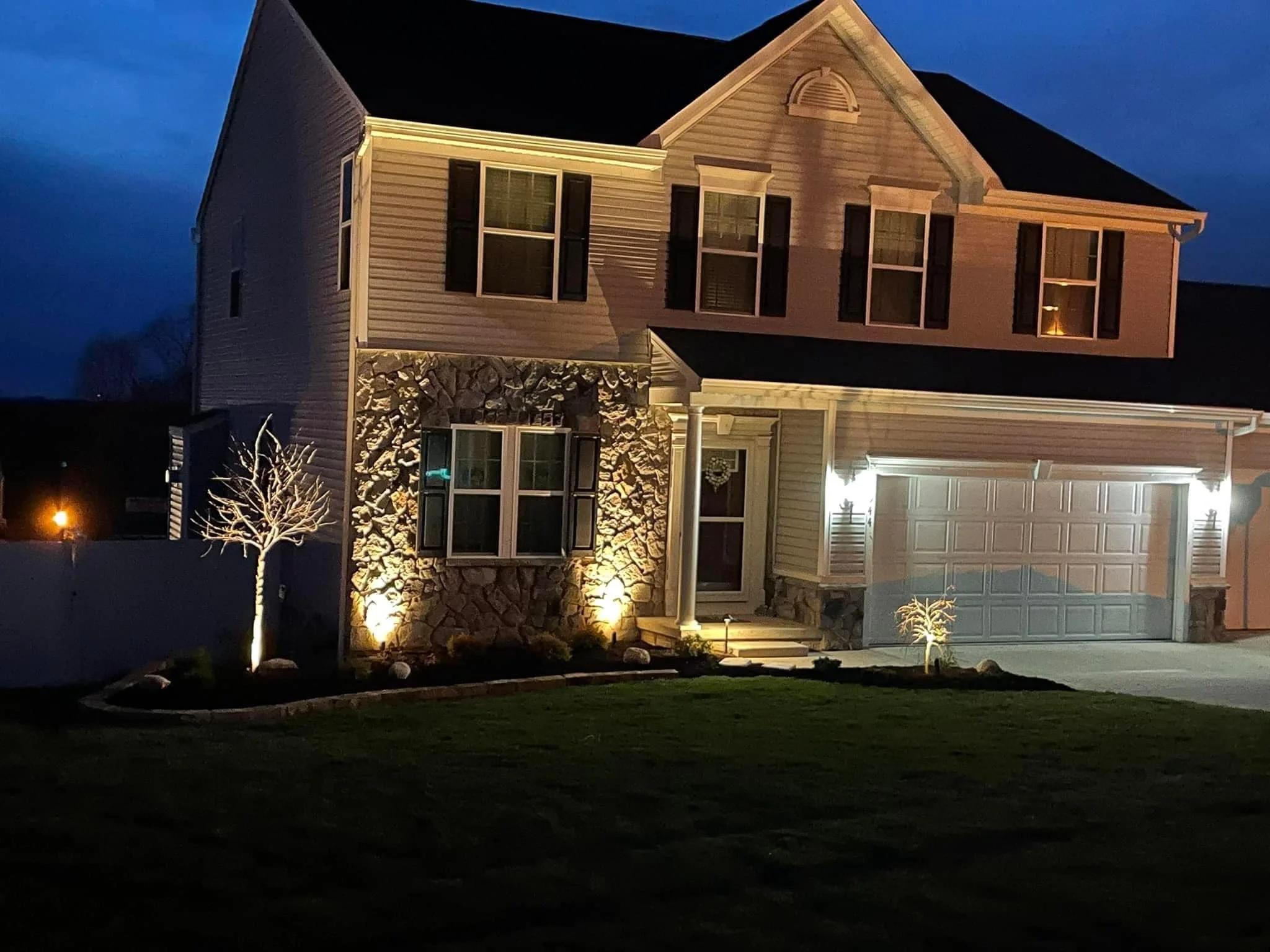 A two-story house illuminated with exterior lighting at night, featuring light-colored siding, dark shutters, a stone accent wall, and an attached garage with closed doors. Small trees with uplighting are in the front yard.