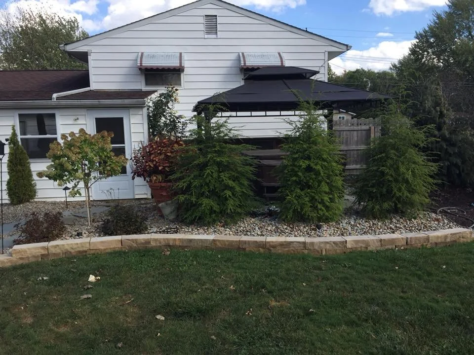 Backyard with a white house, small trees, bushes, gravel area, and a black patio umbrella.