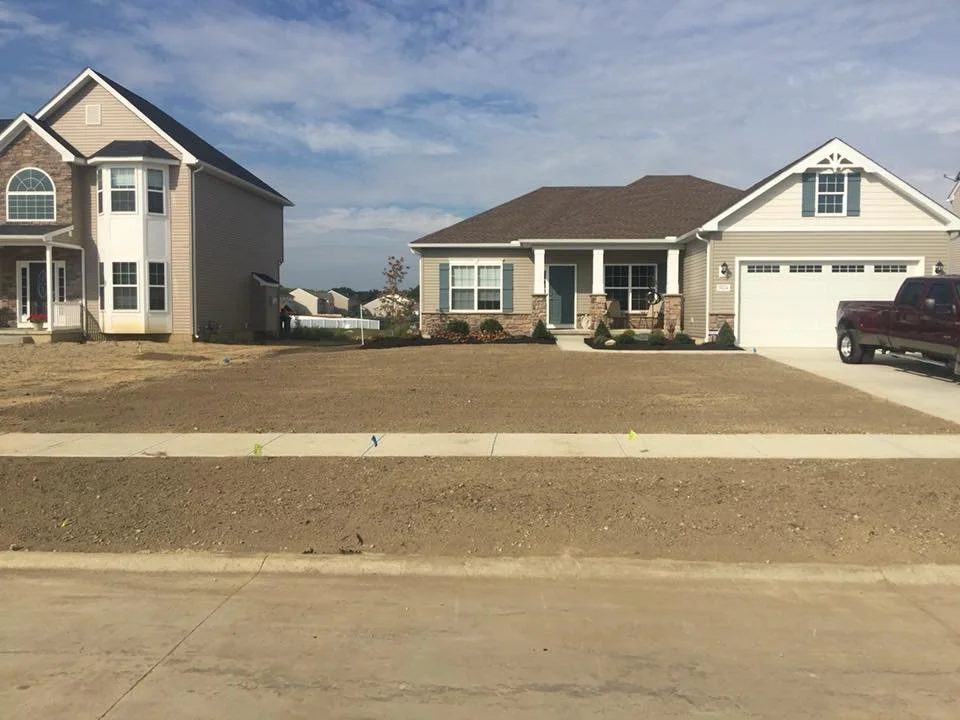 View of two houses with a cleared front yard and sidewalk, one house is under construction with drywall and the other is finished with siding, driveway, and a parked red truck.