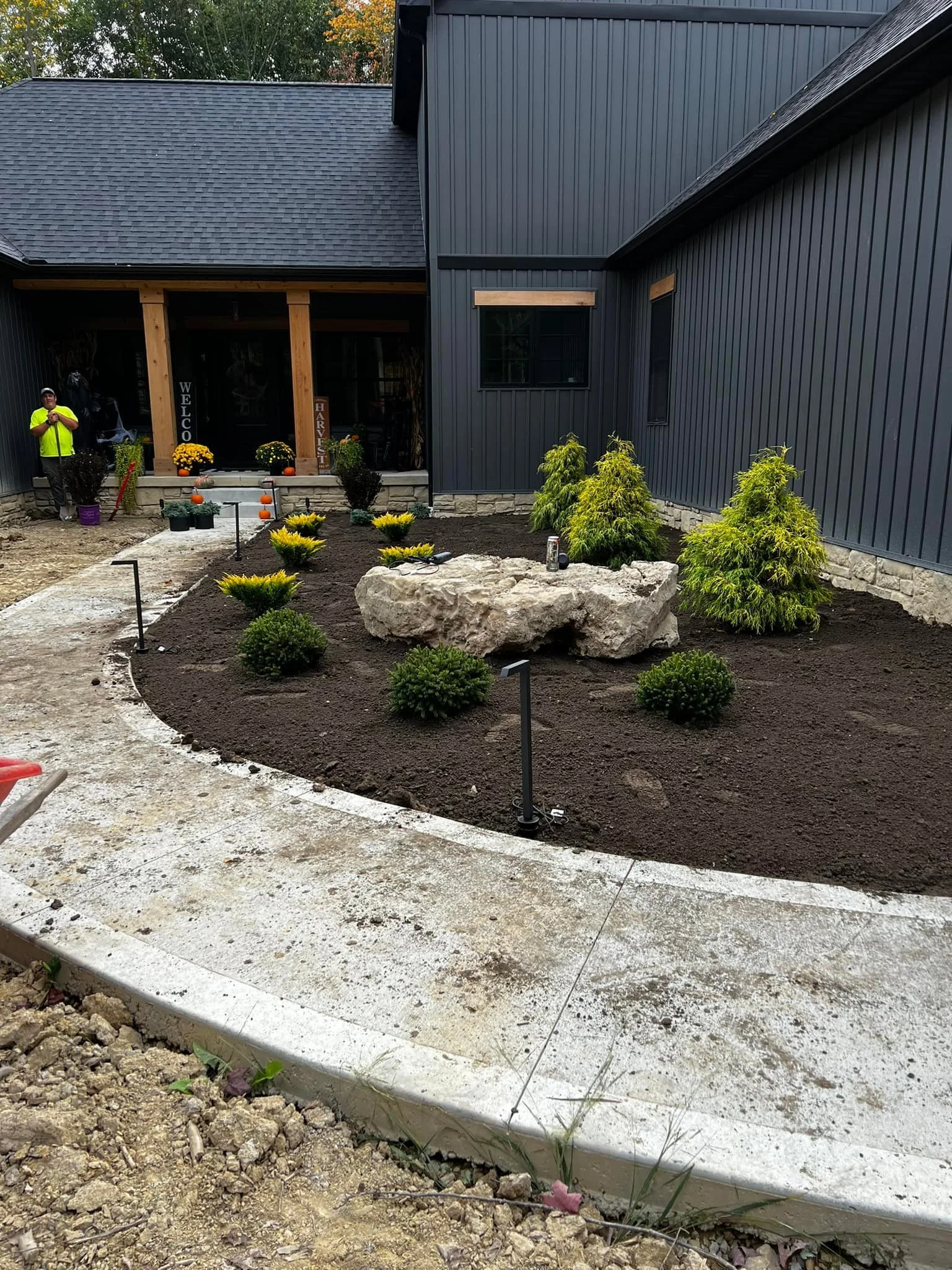 Newly landscaped backyard with small bushes, rocks, and a concrete pathway, next to a house with dark blue siding and large windows.