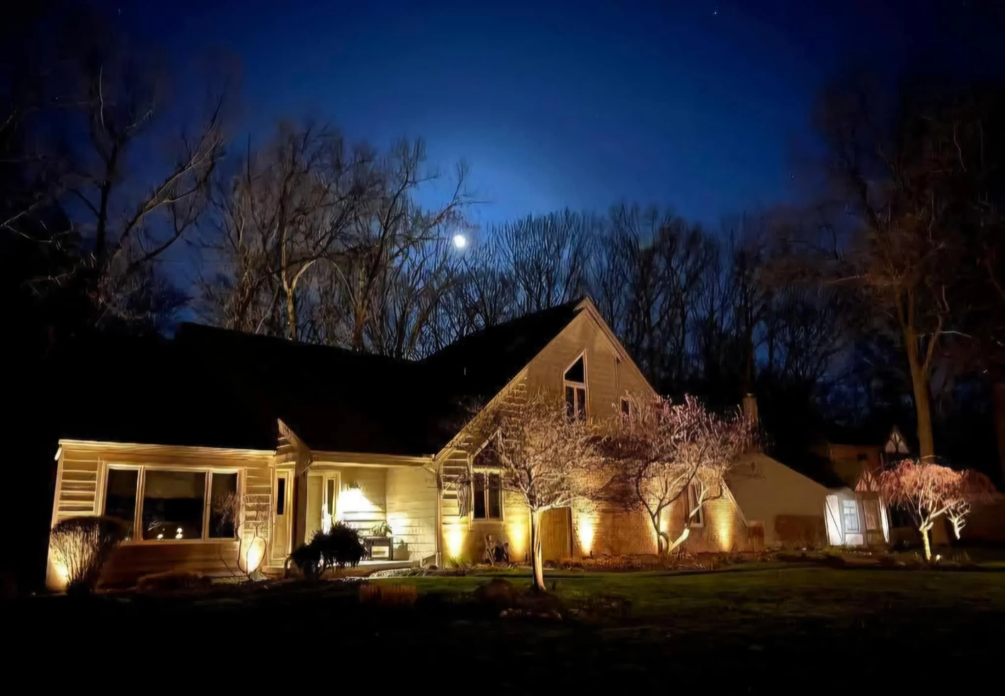 Nighttime view of a house illuminated with exterior lights, trees in the yard, a bright moon in the sky, and a dark blue sky with clouds.