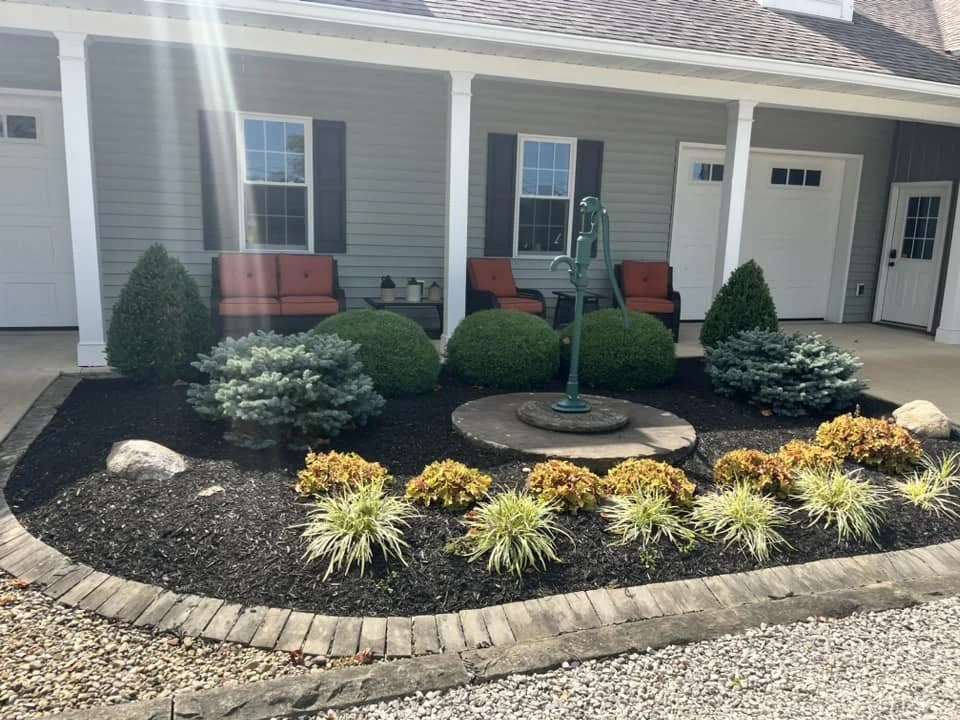Front yard garden with shrubs, plants, and a decorative water pump in front of a house with a porch, two orange chairs, and a closed garage door.