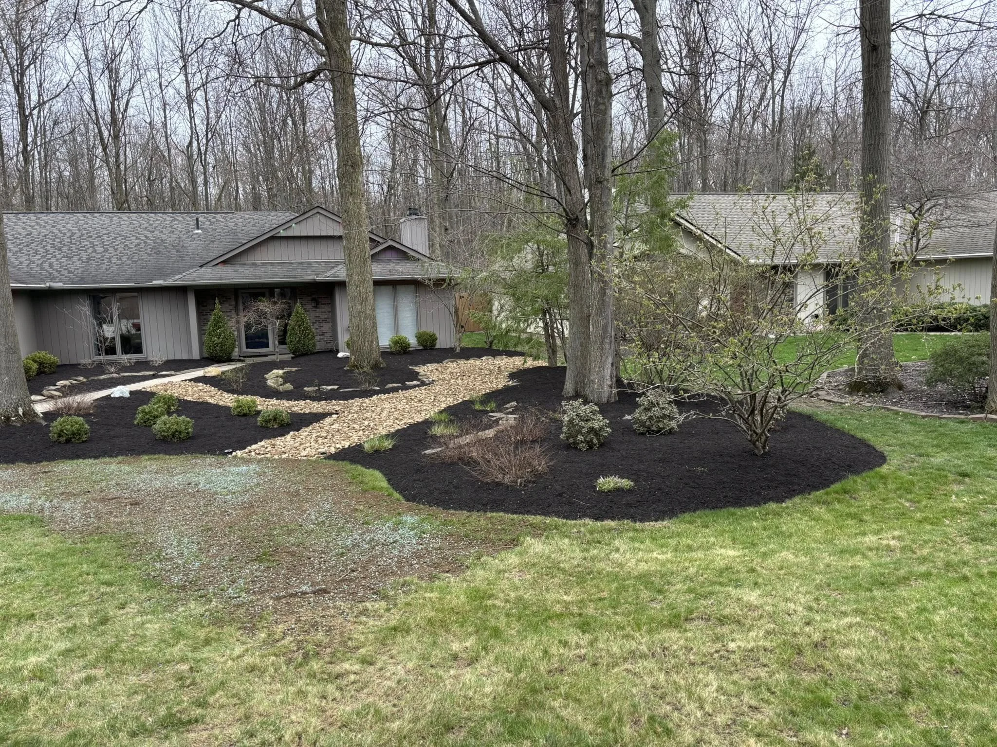 A landscaped backyard with a pathway of white rocks, shrubs, and trees, surrounding a house with a gray exterior and a forested area in the background.
