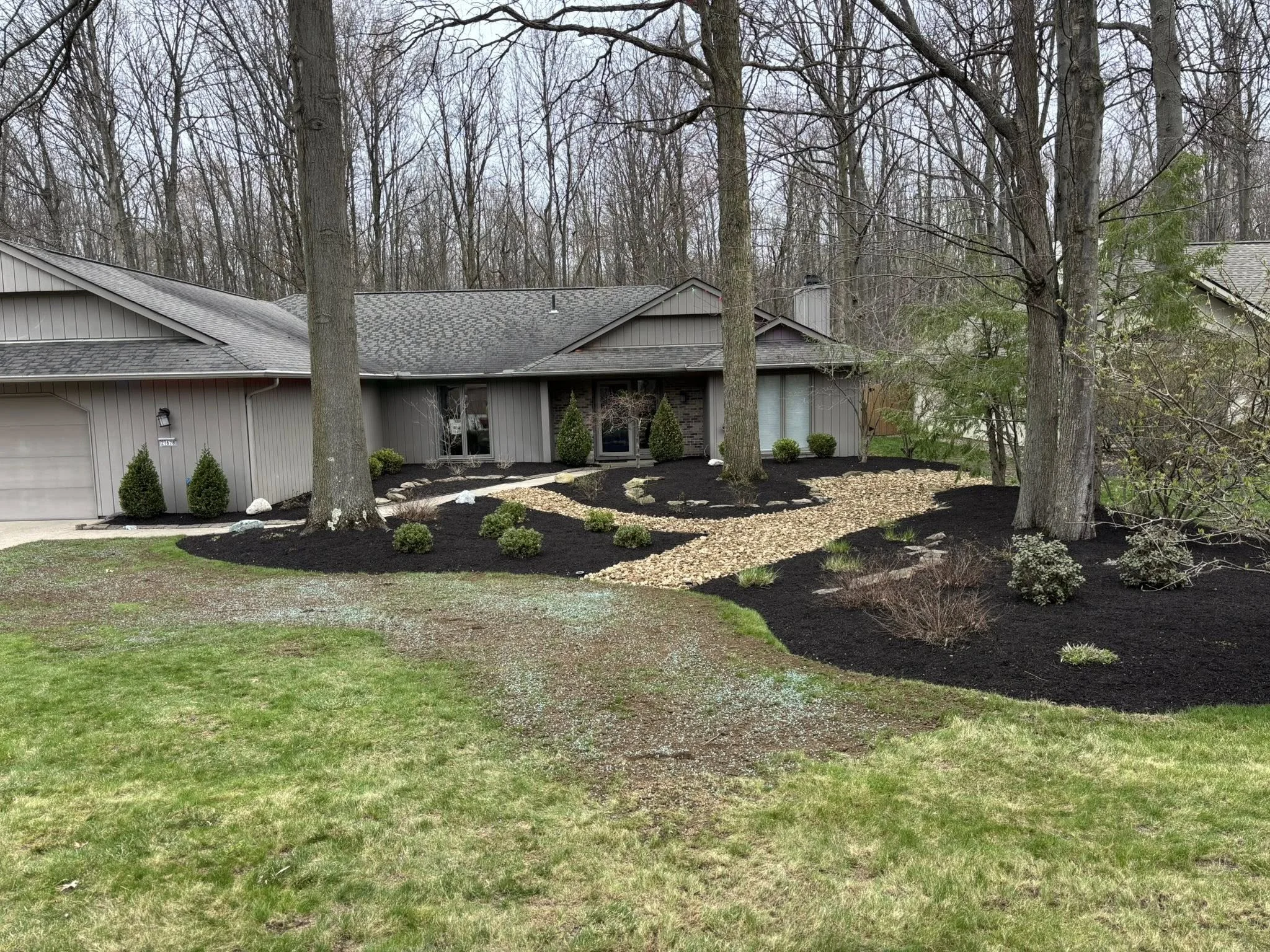 Front yard of a house with freshly landscaped garden beds, mulch, small shrubs, and a winding stone pathway, with large trees and a wooded background.