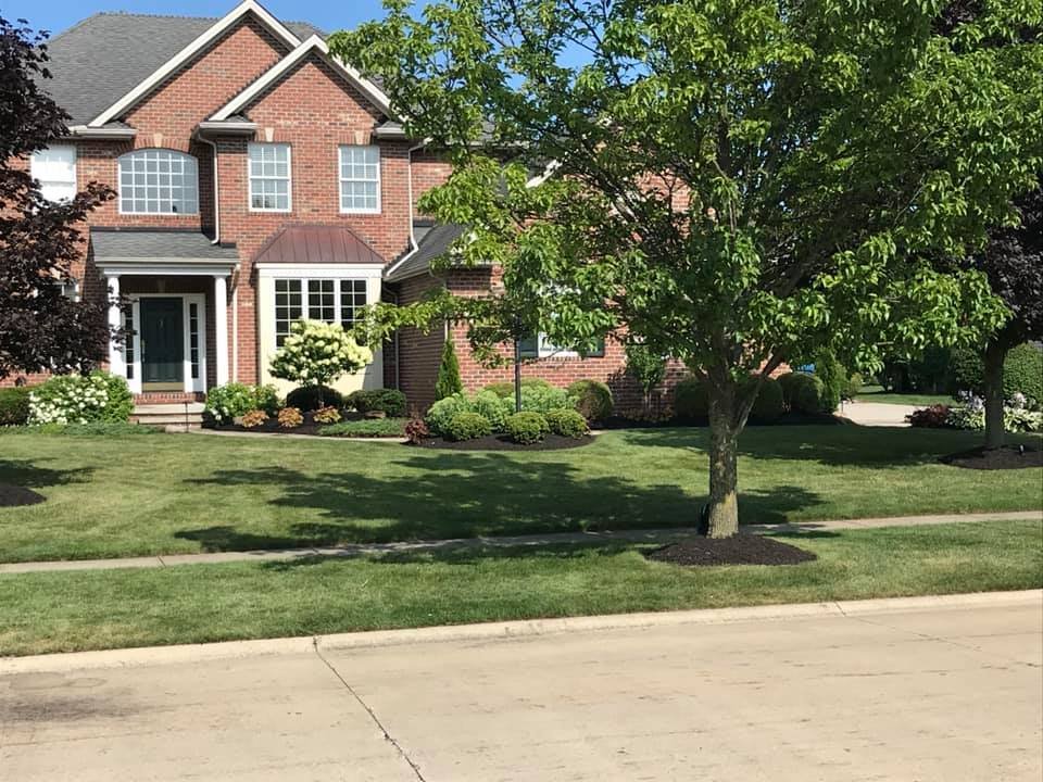 A two-story brick house with a manicured lawn and landscaped garden, including trees, bushes, and flower beds.