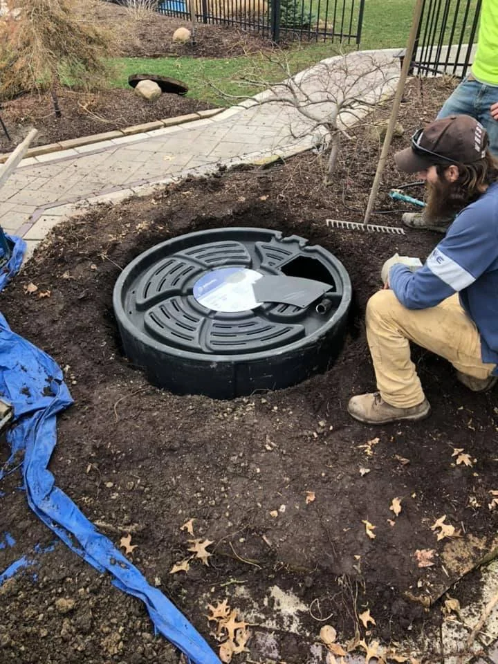 A person kneeling next to a newly installed fountain drain in a backyard with dirt and fallen leaves, with a paved walkway, a small tree, and a fence in the background.