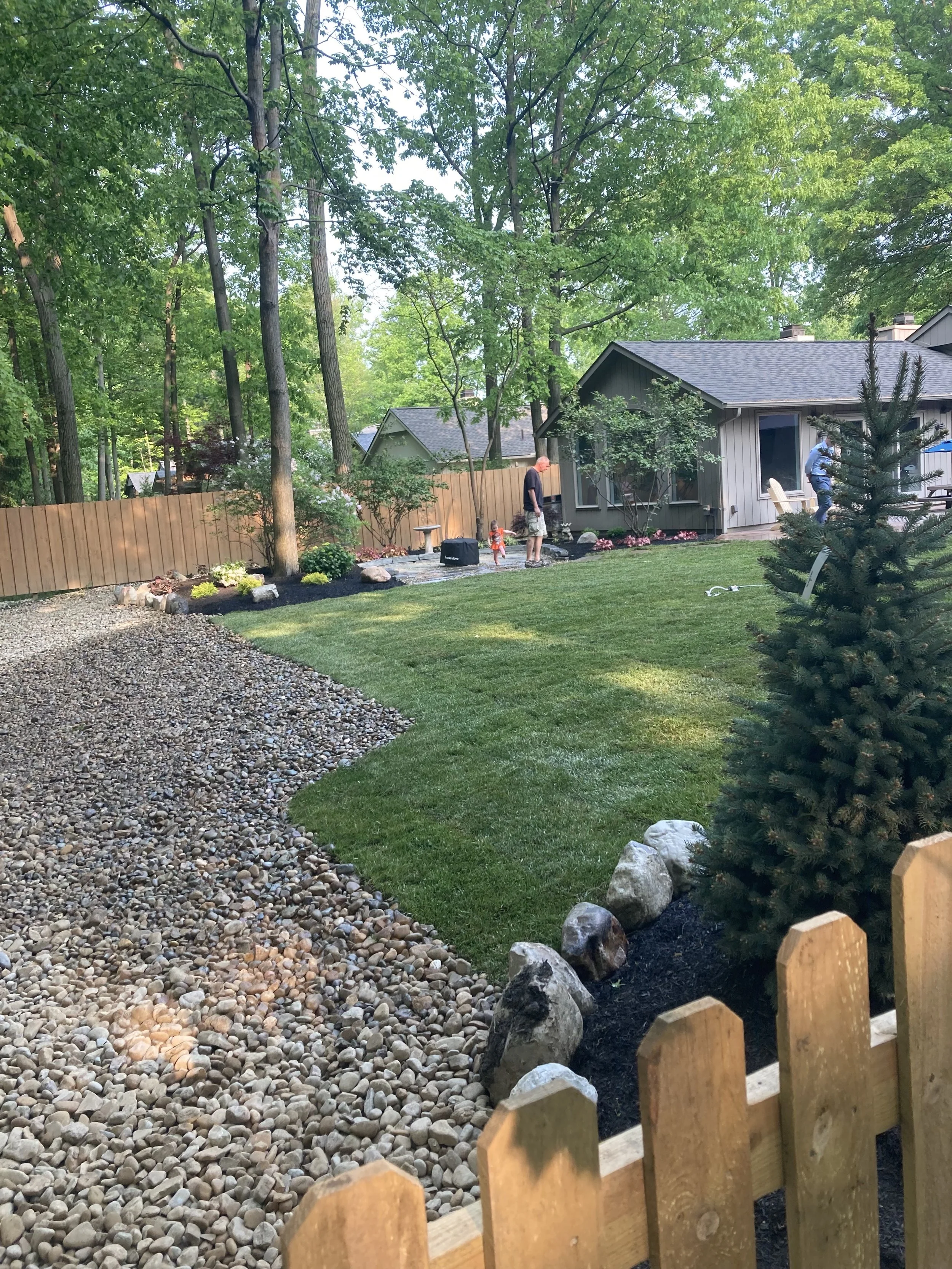 A backyard with a manicured lawn, a stone pathway, trees, a wooden fence, and a house. Two adults, a child, and a dog are visible, with one adult standing near the house, another walking near the trees, and the child on the pathway, all enjoying the 