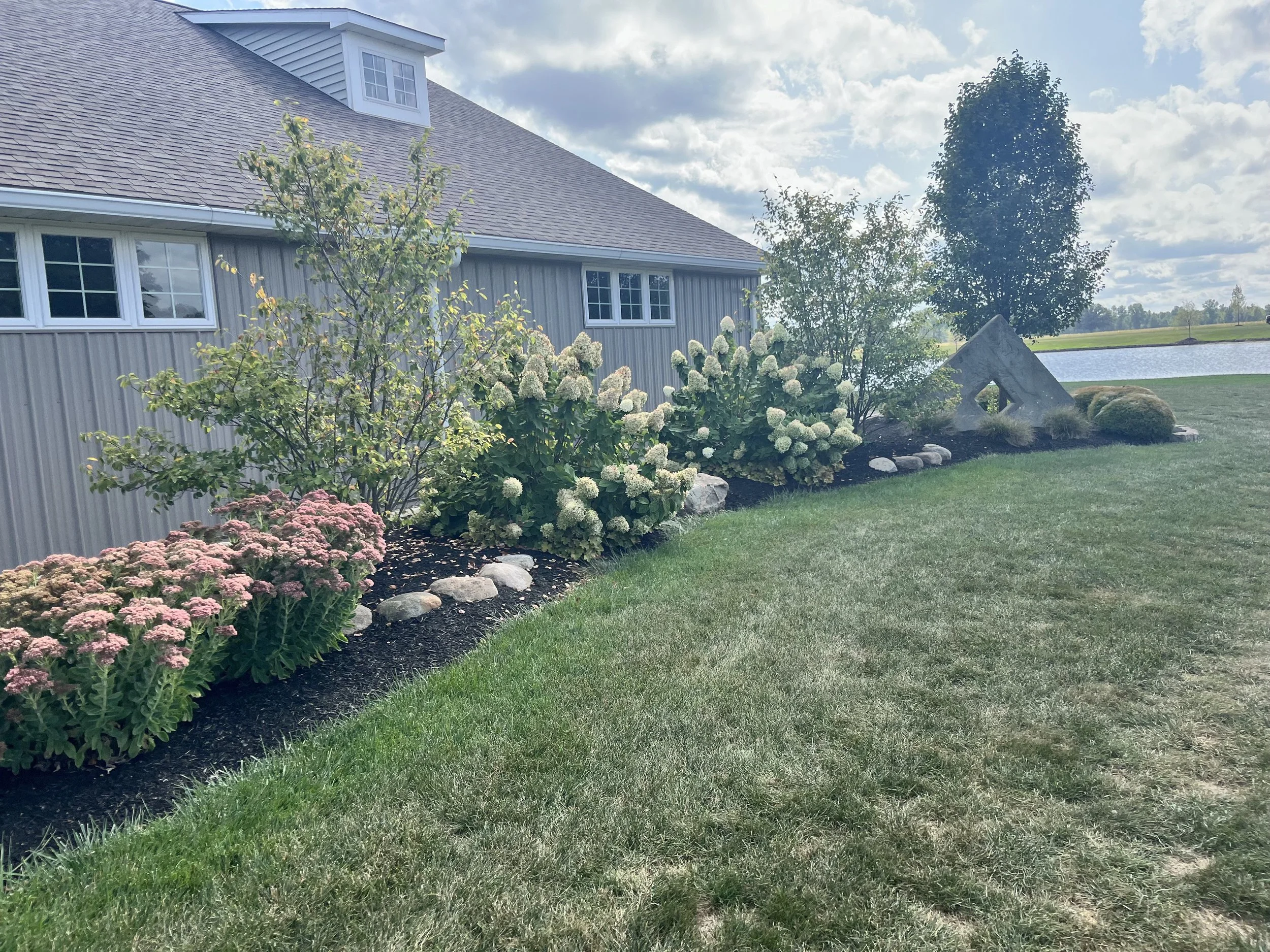 A landscaped yard beside a house with gray siding, featuring pink and white flowering plants, small trees, rocks, and a grassy lawn, with a pond and open sky in the background.
