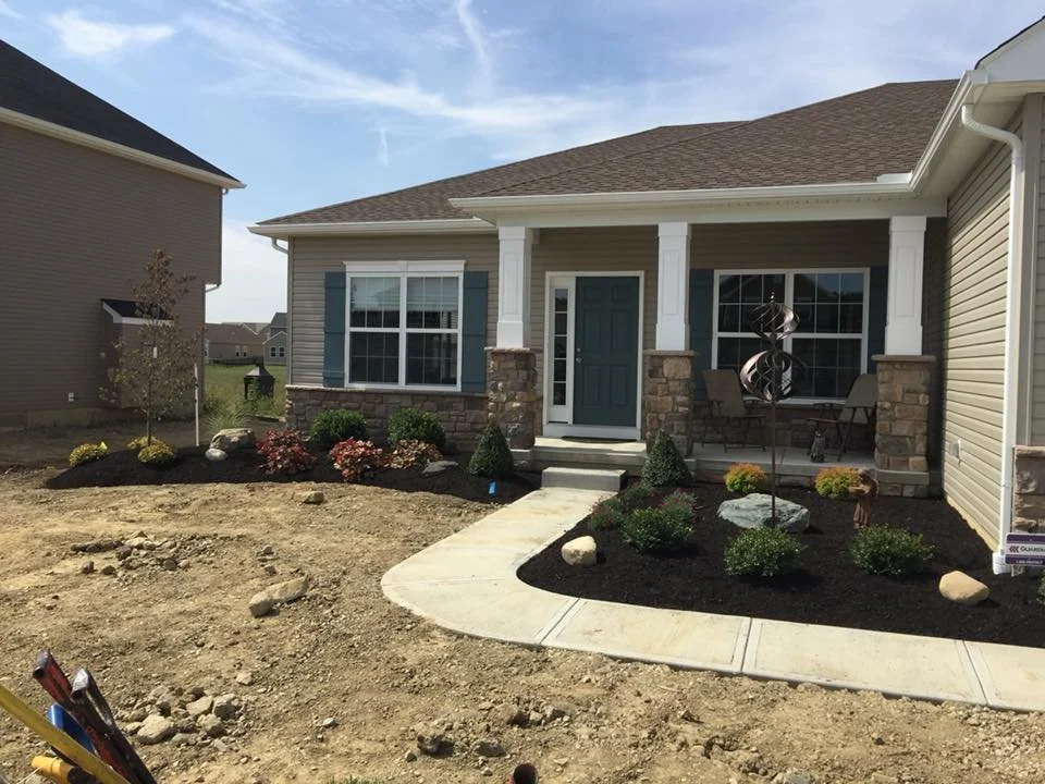 Newly landscaped front yard of a house with small bushes, decorative rocks, and a curved concrete walkway leading to the front door, which is painted dark blue. The house has blue shutters, stone accents, and a porch with two chairs. The yard is stil