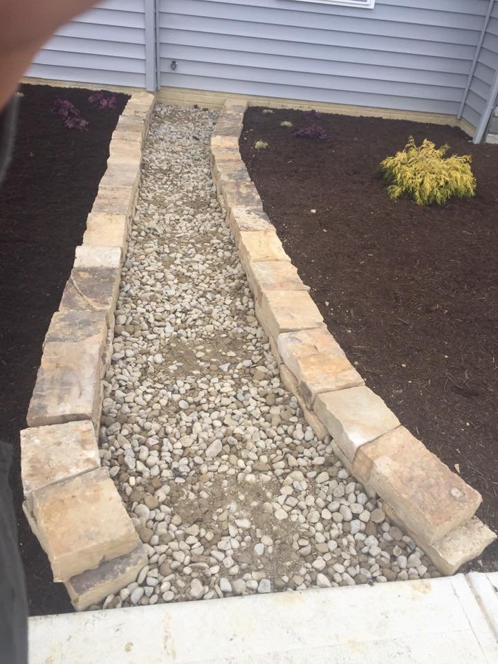 A newly constructed gravel pathway lined with large stone bricks, adjacent to a flower bed with dark mulch and some small plants, next to a light gray siding wall.