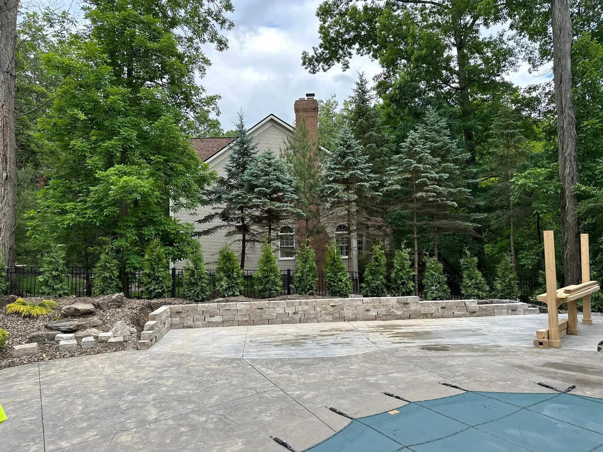 A partially constructed backyard patio with a stone retaining wall, surrounded by tall evergreen trees and a house in the background.