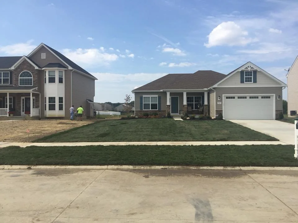 Two houses under construction, with workers in the yard of the left house. The house on the right has a finished lawn and driveway, while the house on the left is still being landscaped.