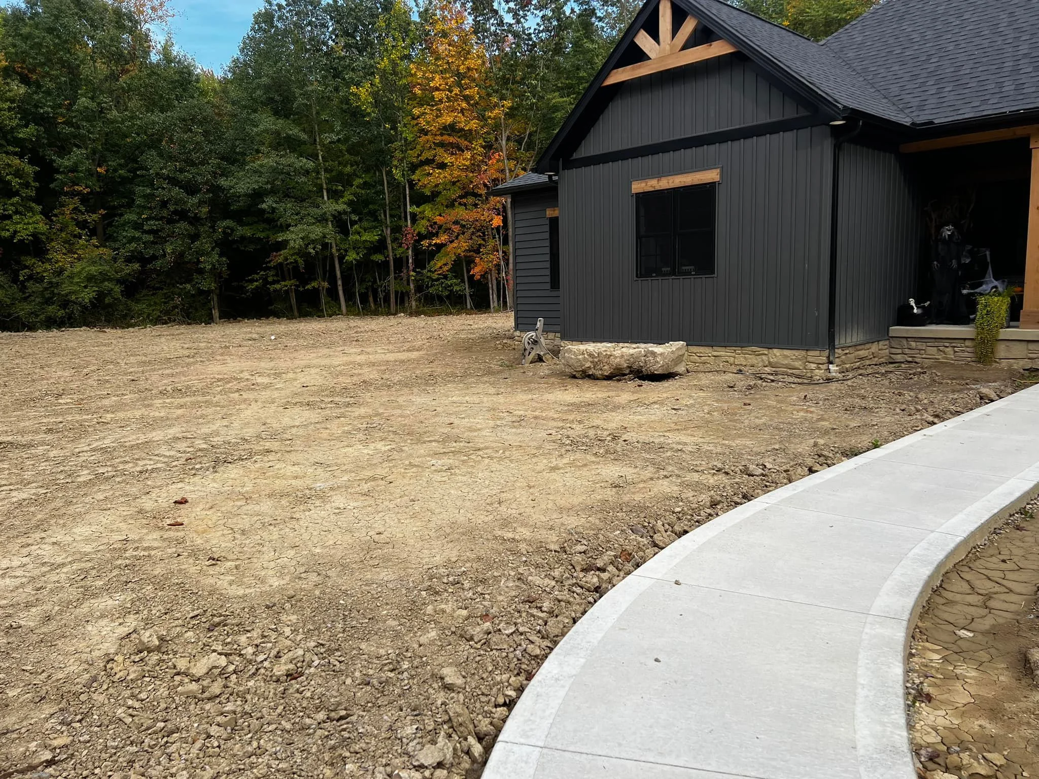 A house with black siding and a gray roof, situated next to a new curved concrete sidewalk. The yard in front of the house is bare dirt, surrounded by trees with some leaves turning color, indicating fall. There is construction equipment and a large 