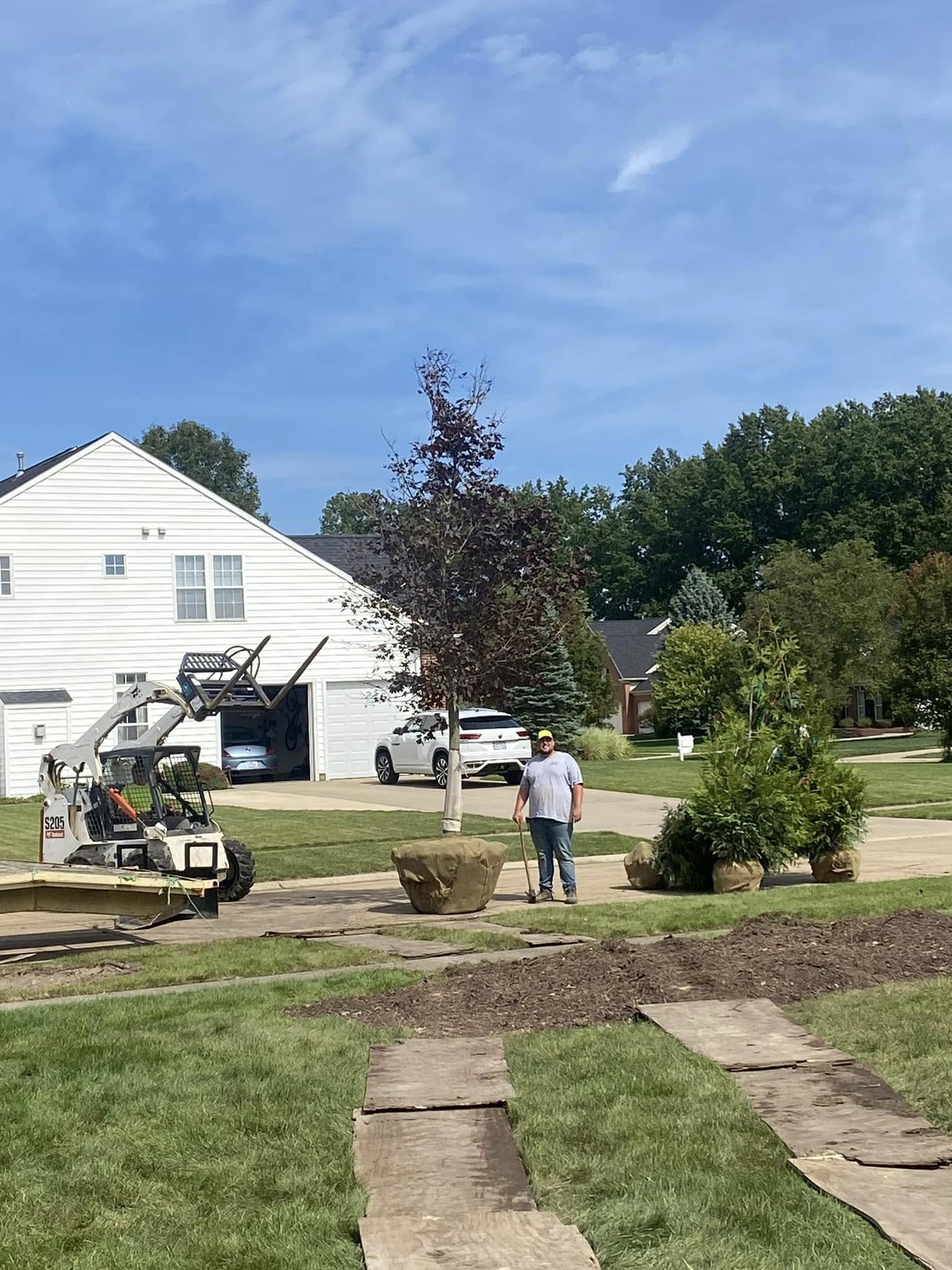 A person standing outdoors near a small tree and large decorative rocks, with construction equipment and a white house in the background.