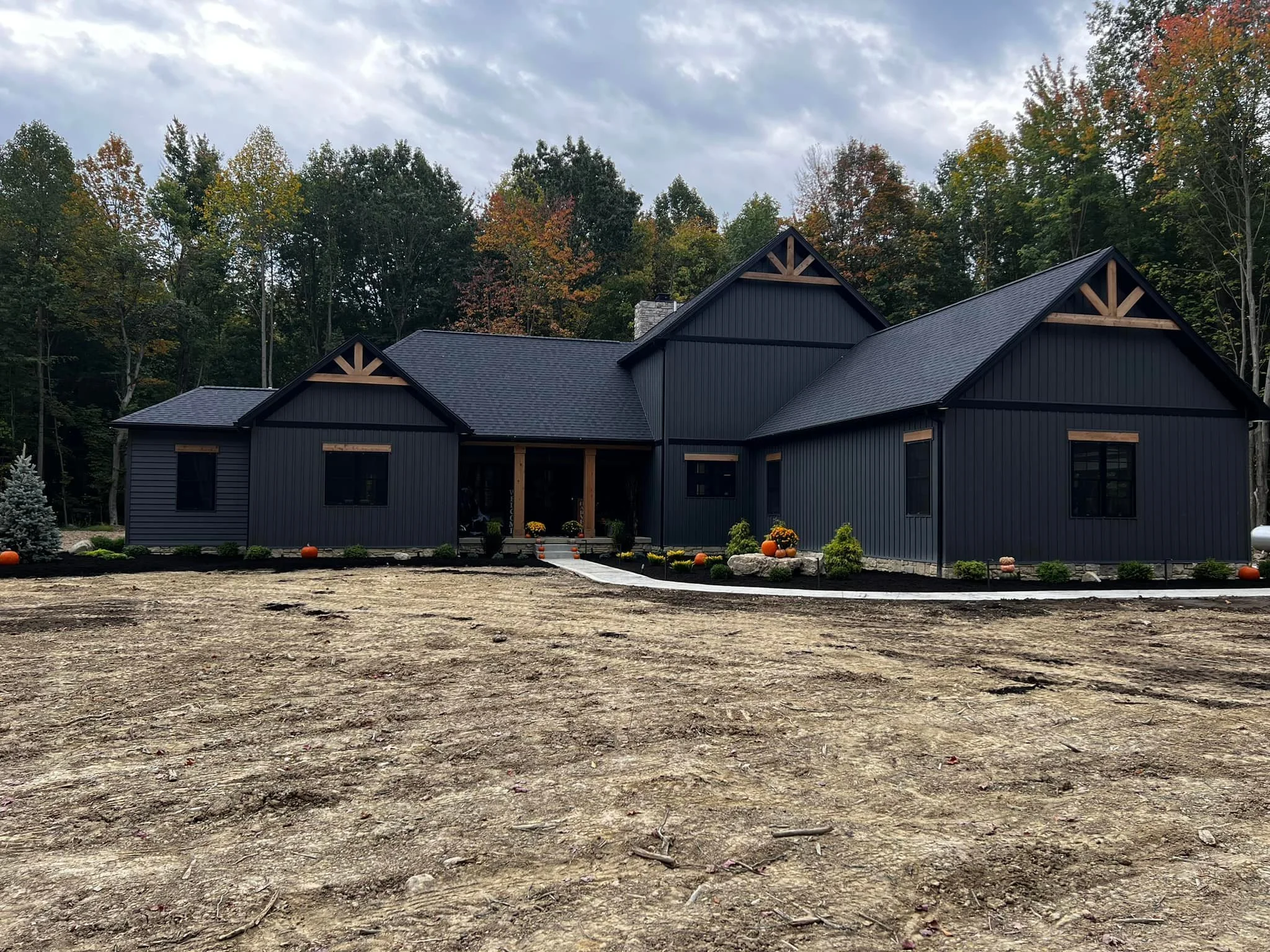 A recently built black house with a gabled roof, surrounded by some autumn trees, featuring a landscaped front yard with pumpkins and potted plants.