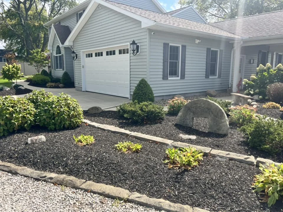 A suburban house with light gray siding, black shutters, and a white garage door with rectangular windows. The front yard has a landscaped garden with mulch, small shrubs, and a large decorative stone, with a paved driveway and pathway.