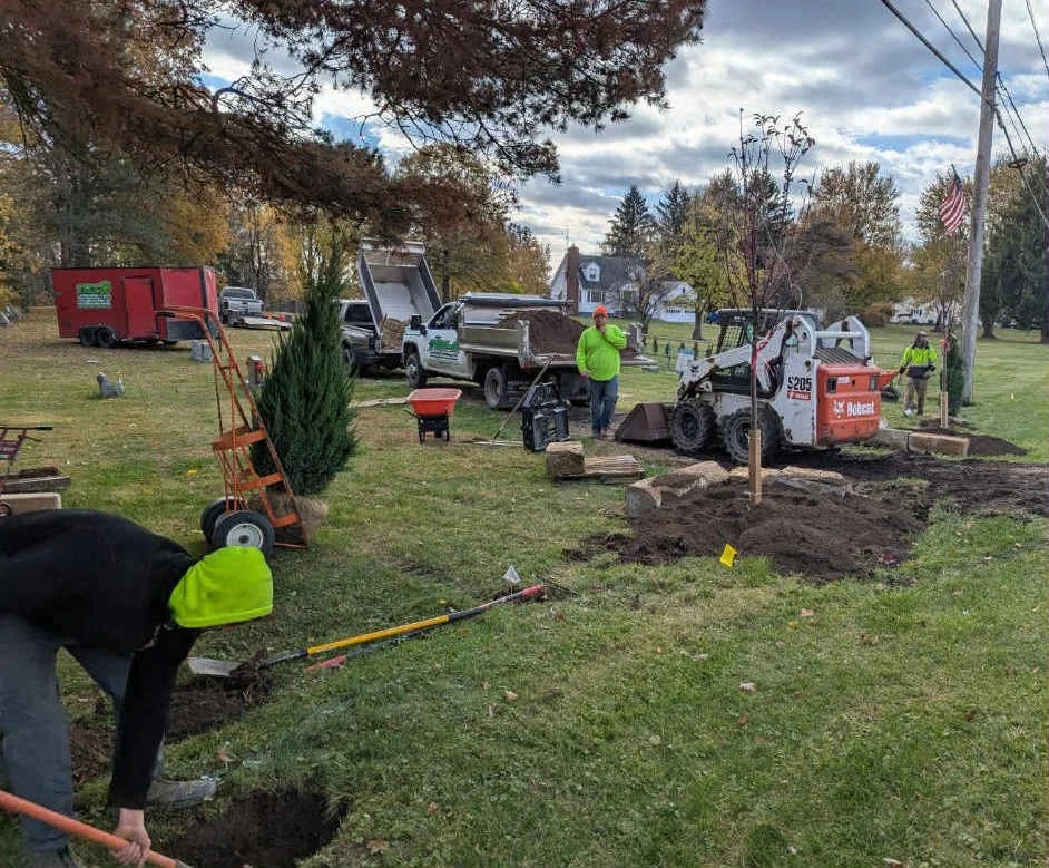Workers are installing landscaping or fencing around a tree in a residential yard. They are using compact machinery and shovels, with several trucks and equipment nearby. The scene is set in autumn, with trees showing fall colors, and an American fla