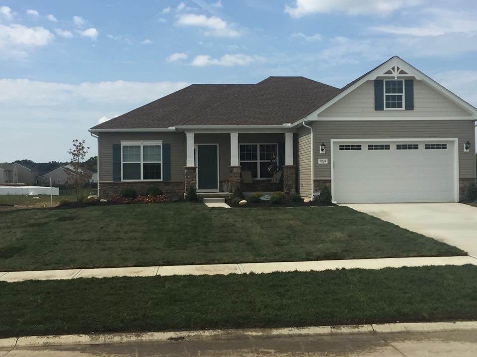 A single-story house with a front lawn, concrete driveway, and attached garage. The house has beige siding, dark grey roof, and white trim with black shutters. There is a small porch with two white columns and a front door.
