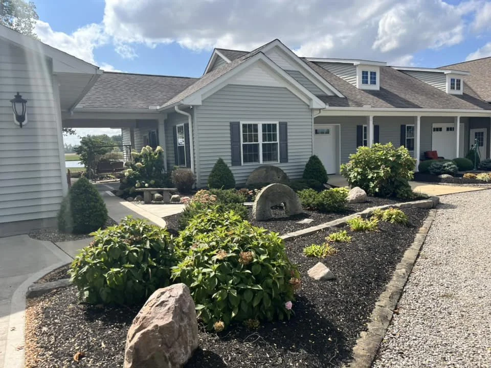 Front yard with neatly landscaped garden beds, bushes, rocks, and large decorative stones in front of a light blue house with white trim and multiple dormer windows.