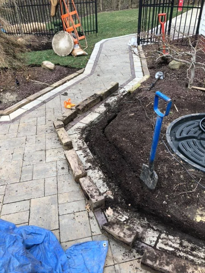 In the process of repairing or constructing a brick walkway in a backyard, with tools and bricks scattered around, a partially completed pathway with a curved design is visible, along with a blue tarp and gardening tools.