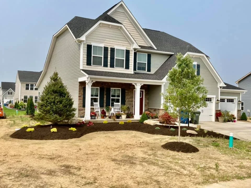 A two-story suburban house with a front porch, surrounded by a well-maintained yard with newly planted grass, trees, and shrubs.