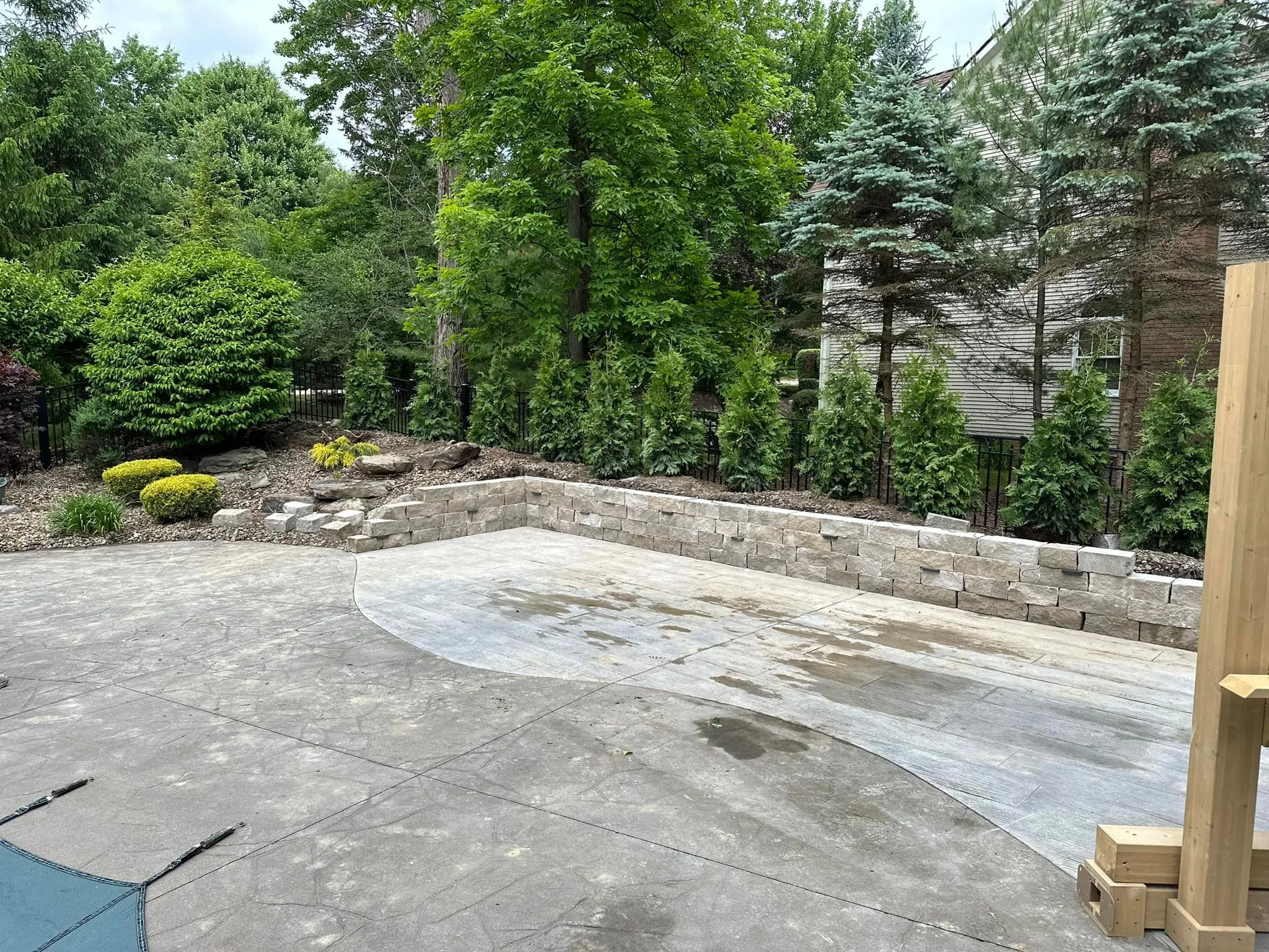 Backyard with a concrete patio, a brand new brick retaining wall, lush green trees and bushes, and a house in the background. There are small rocks and mulch in the garden bed, and part of a wooden post is visible on the right.